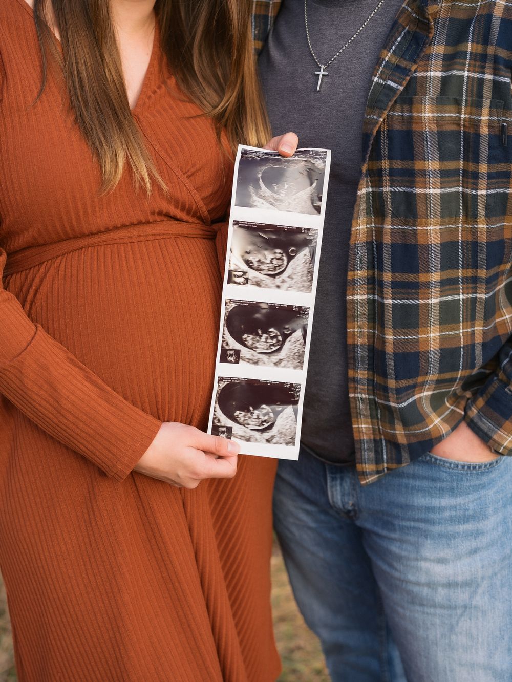 Pregnant couple holding ultrasound photos outdoors, woman in rust wrap dress cradling baby bump.