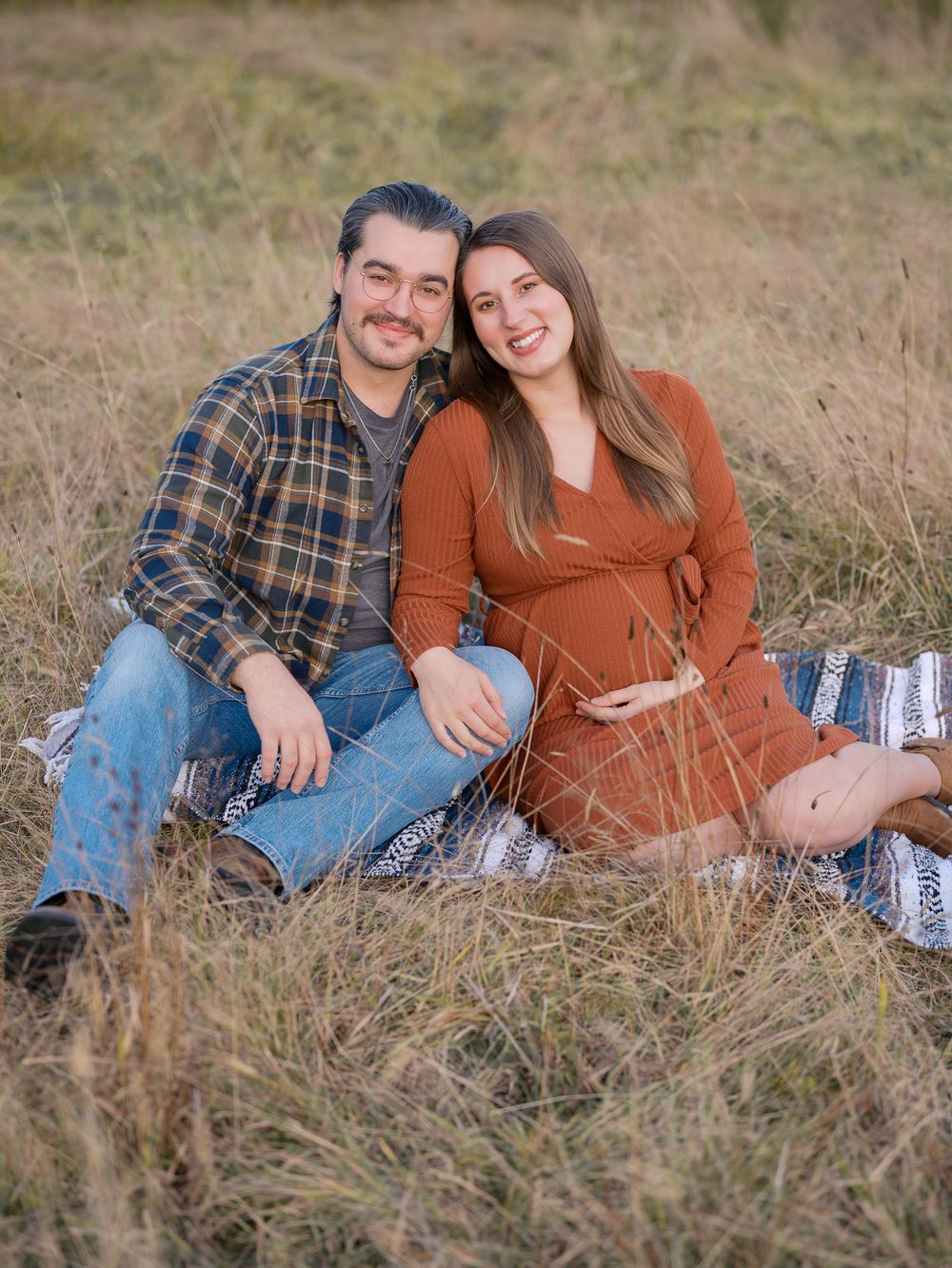 A couple sits together in a grassy field, the woman in a rust dress cradling her baby bump during a maternity photo session.