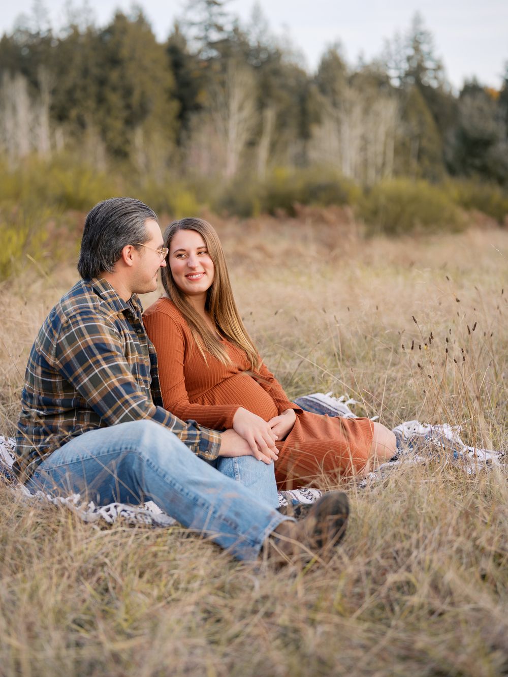 Couple sitting on blanket in golden field, woman in rust dress cradling baby bump, man in plaid shirt, trees in background.