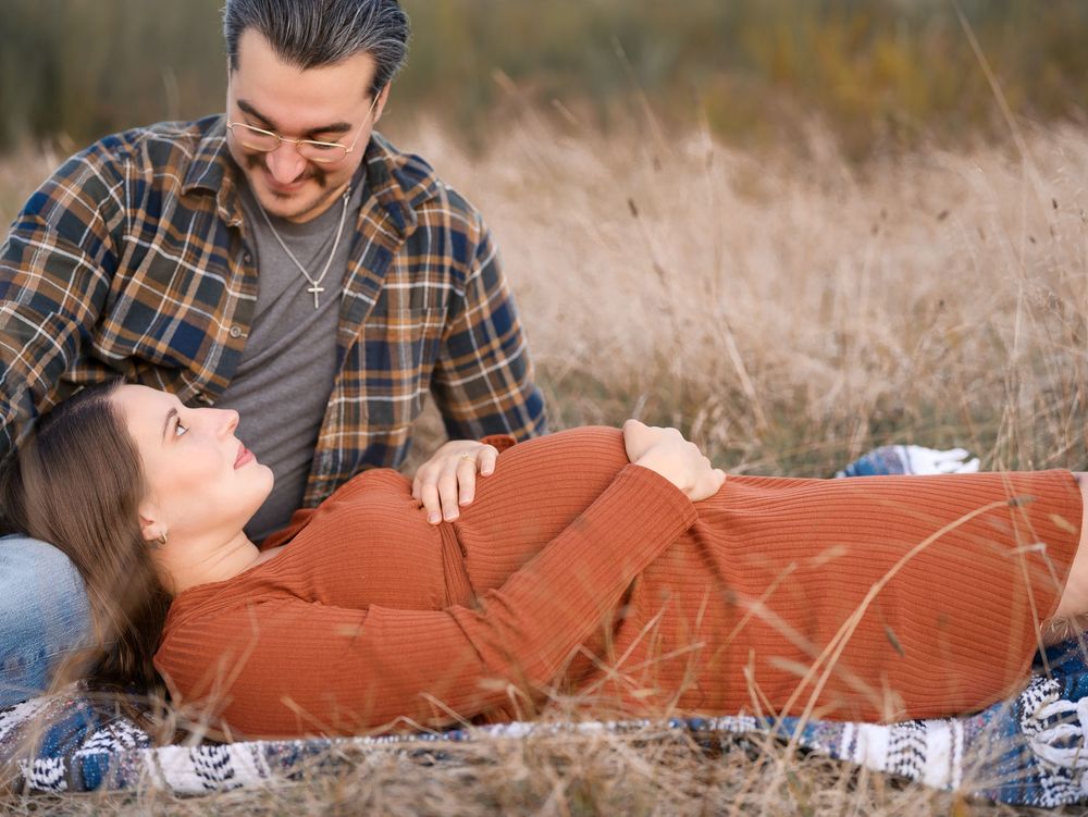 Smiling couple sharing tender moment in autumn field, pregnant woman in rust dress lying down.