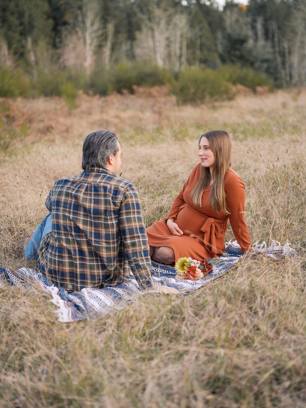 Expecting couple sitting on blanket in golden field having conversation with fall flowers nearby.