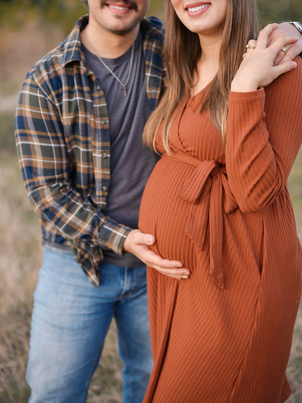 Close-up of couple cradling pregnant belly outdoors, woman in ribbed rust maternity wrap dress.