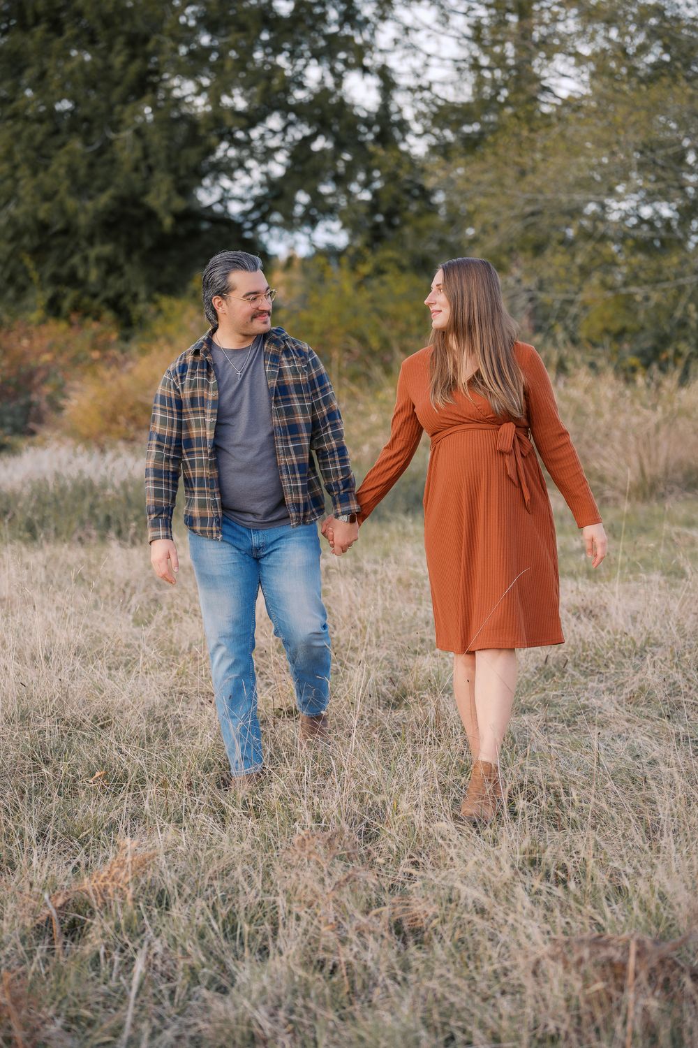 Happy expecting couple walking hand-in-hand through autumn meadow, woman in rust maternity dress.