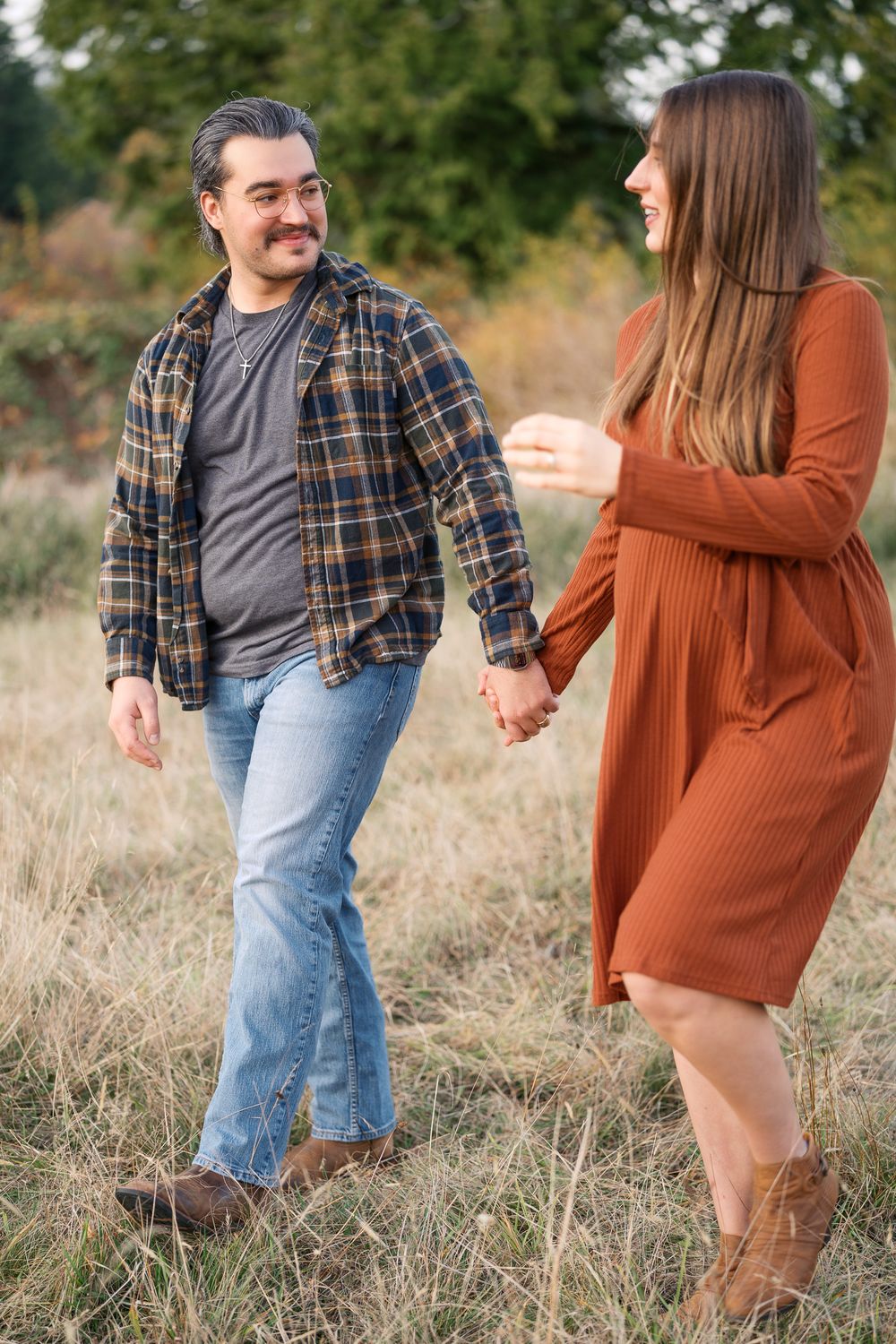 A couple holds hands and smiles while walking through a field in autumn, dressed in casual fall outfits.
