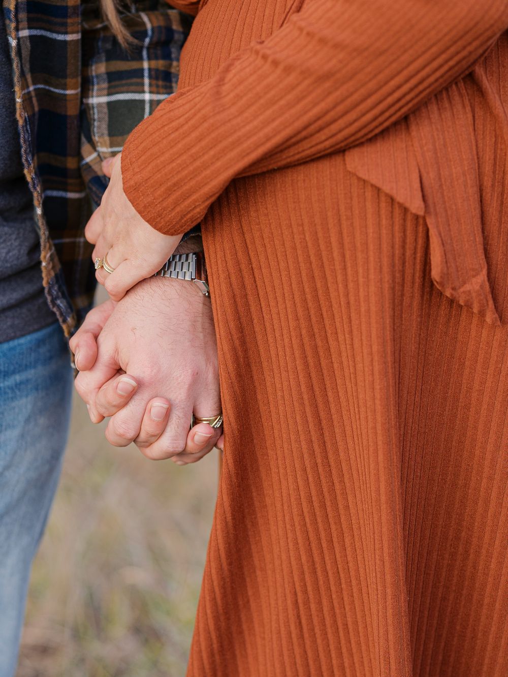 A couple holds hands tenderly, wearing wedding rings, dressed in an orange ribbed dress and plaid flannel shirt outdoors.