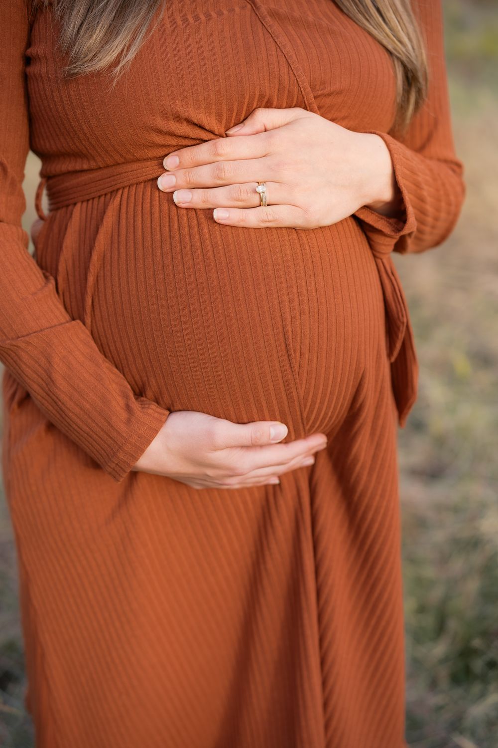 Pregnant woman in rust ribbed wrap dress cradling baby bump with both hands, wearing gold ring.