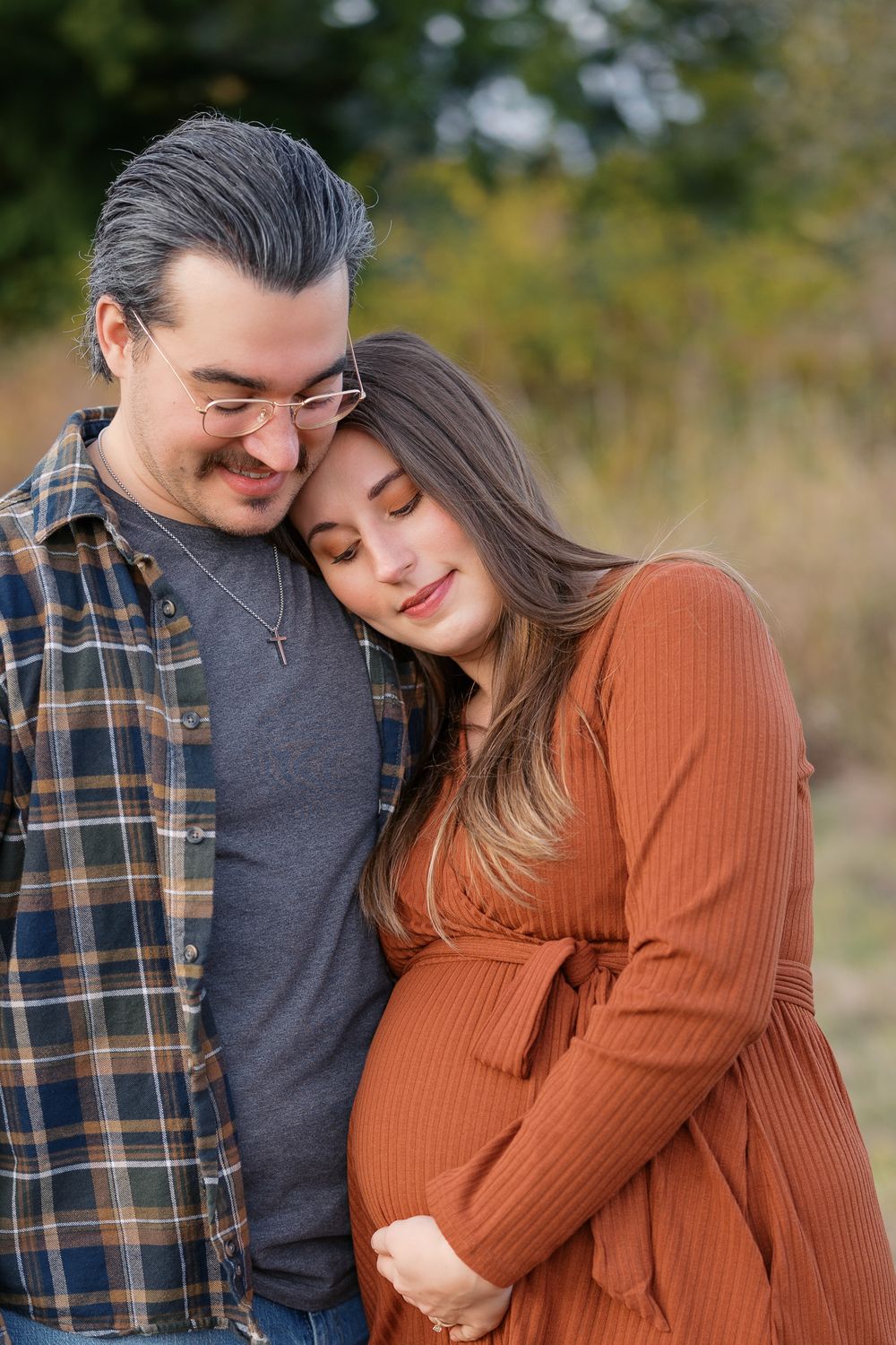 A couple shares a tender moment outdoors, the pregnant woman in rust dress leaning on her partner during a maternity photo session.