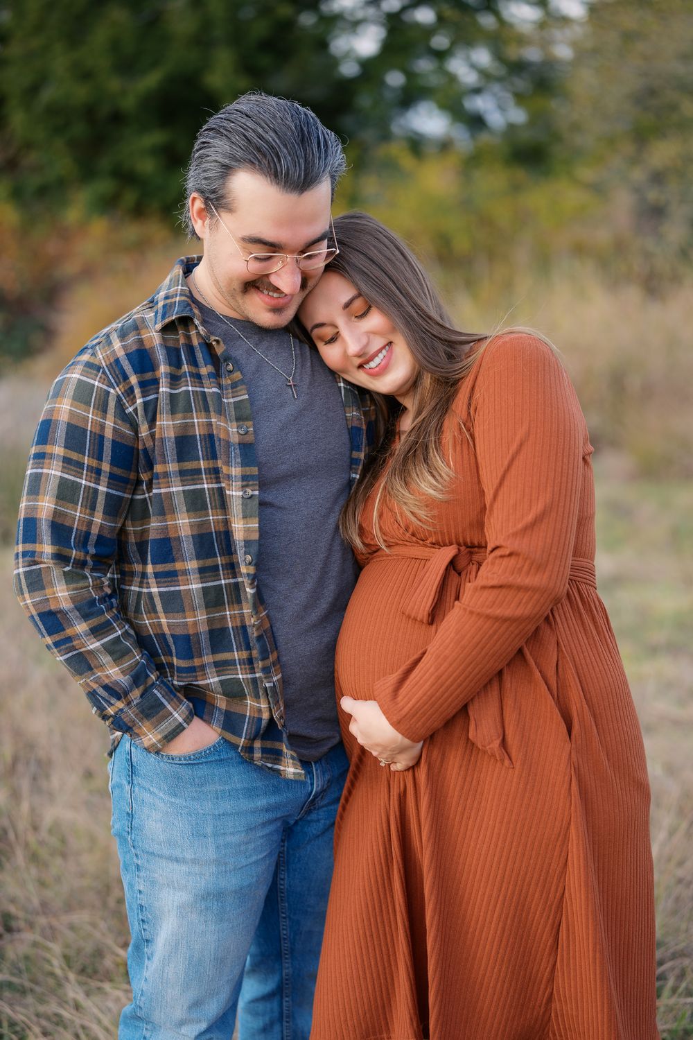 Expecting couple embracing tenderly in fall outdoor setting, woman in rust dress cradling baby bump.