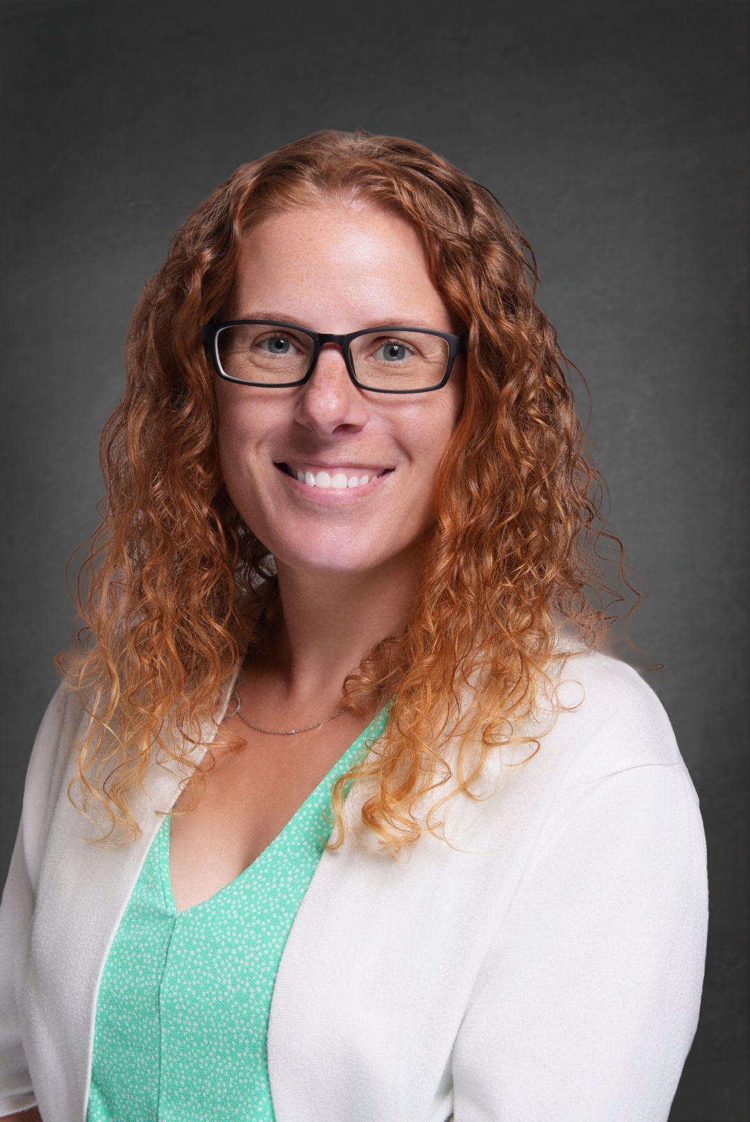 Professional headshot of a smiling woman with curly red hair and glasses wearing a white cardigan over a mint green top.