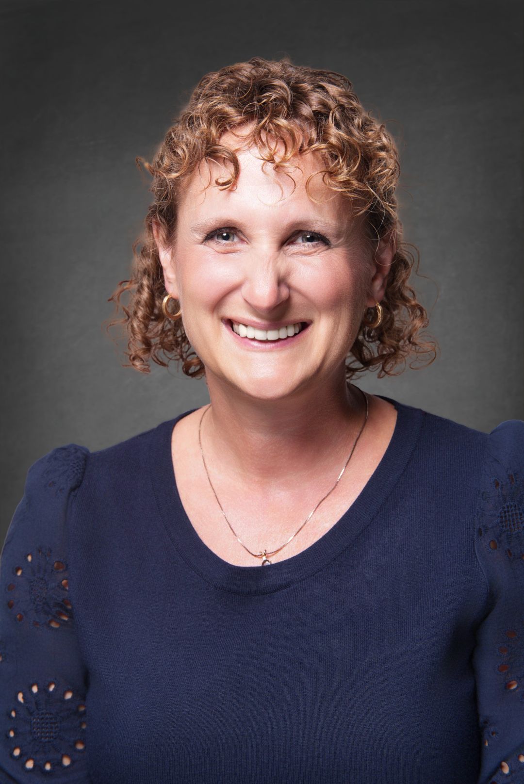 Smiling woman with curly auburn hair, hoop earrings and a gold necklace wearing a navy embroidered top in a professional headshot.