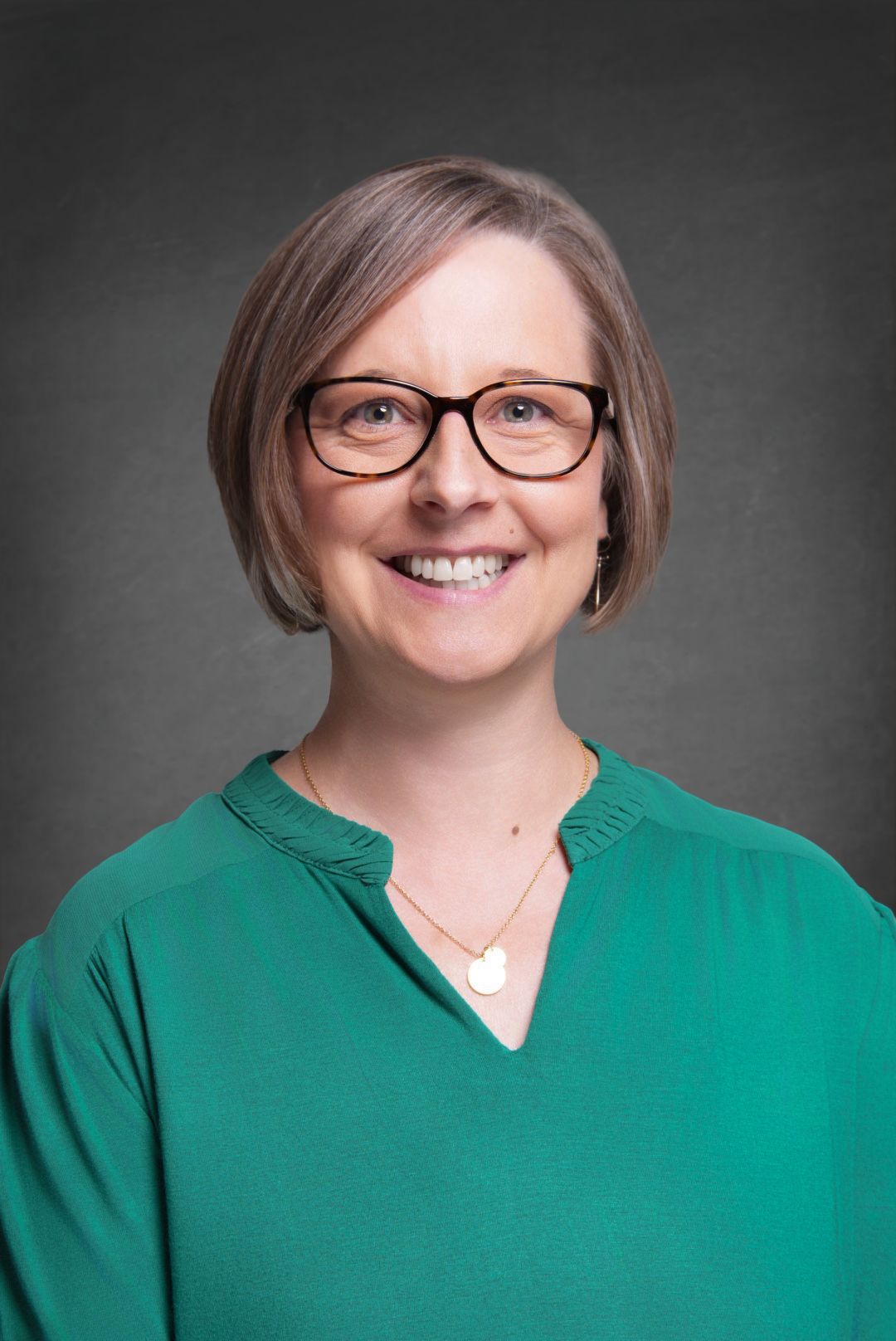 Smiling woman with a short brown bob and dark-rimmed glasses wearing a green blouse and a round pendant necklace in a professional headshot.