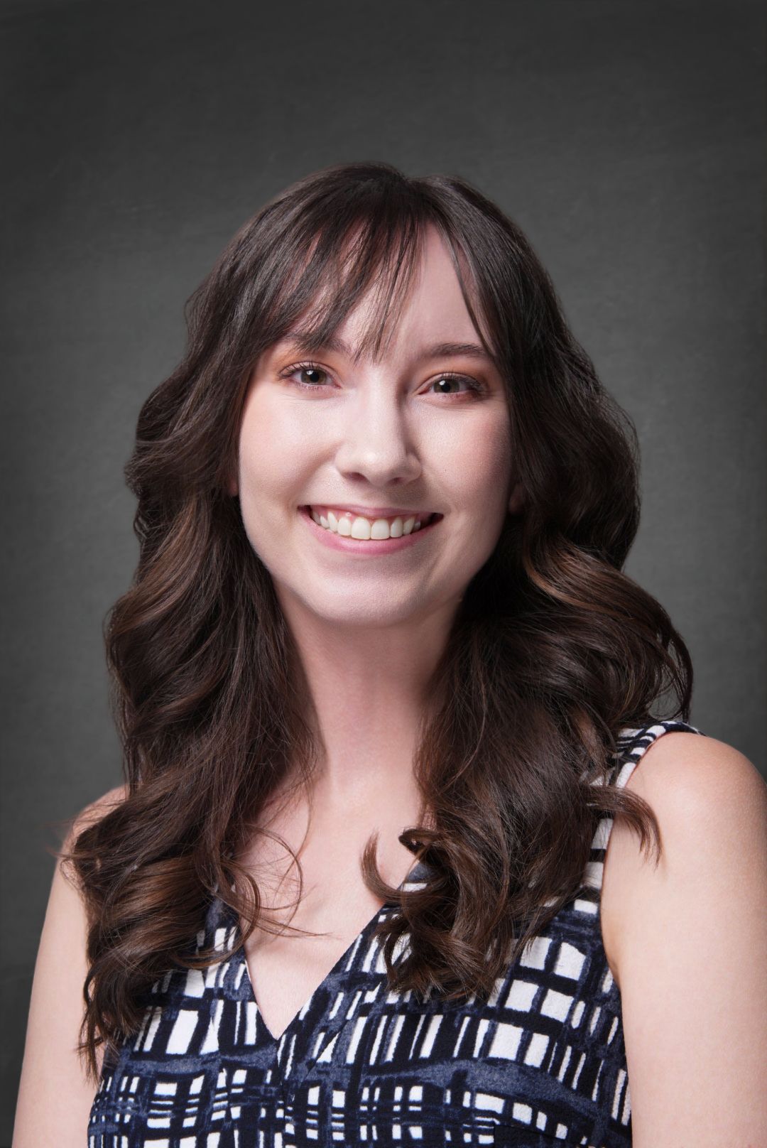 Young woman with long wavy brown hair and bangs smiling in a navy and white patterned sleeveless top against a gray studio background.