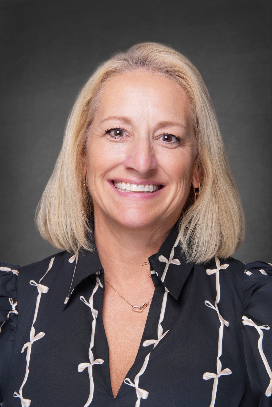 Smiling woman with shoulder-length blonde hair wearing a black collared blouse with white bow pattern and a delicate necklace in a headshot.