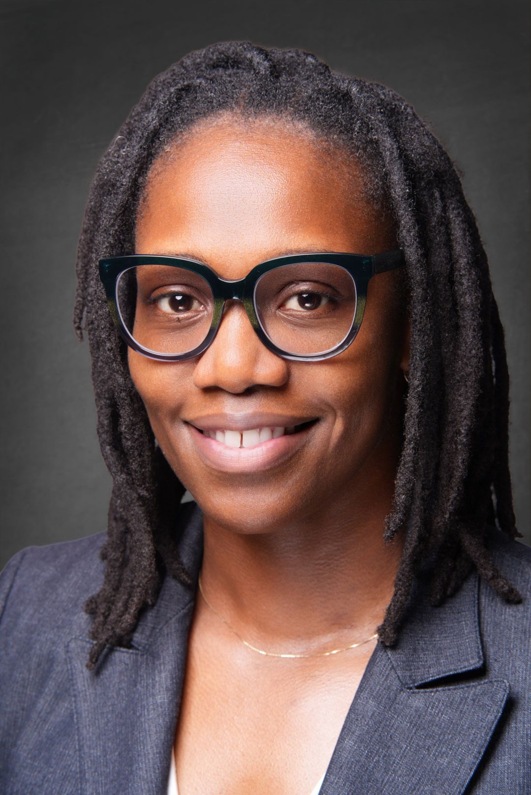 Smiling woman with long locs and bold dark green glasses wearing a gray blazer and a thin gold necklace in a professional portrait.