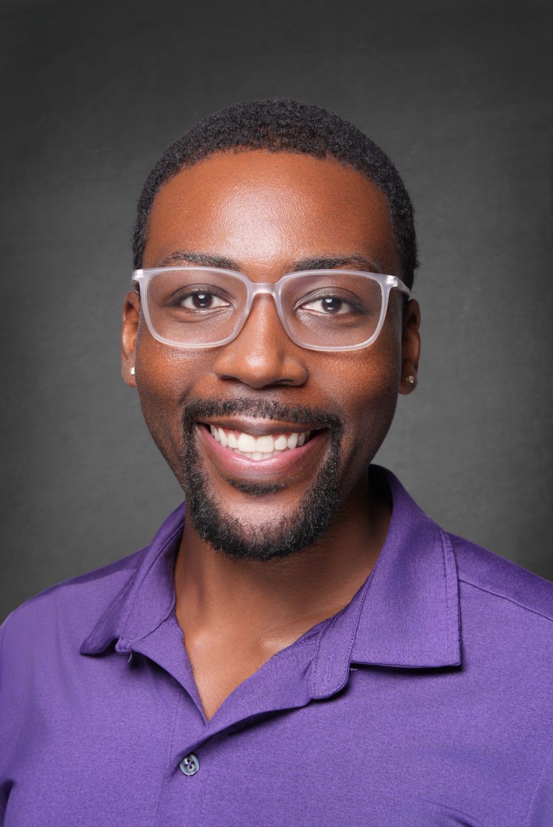 Smiling man with a trimmed beard and clear-framed glasses wearing a purple polo shirt in a professional headshot against a gray background.