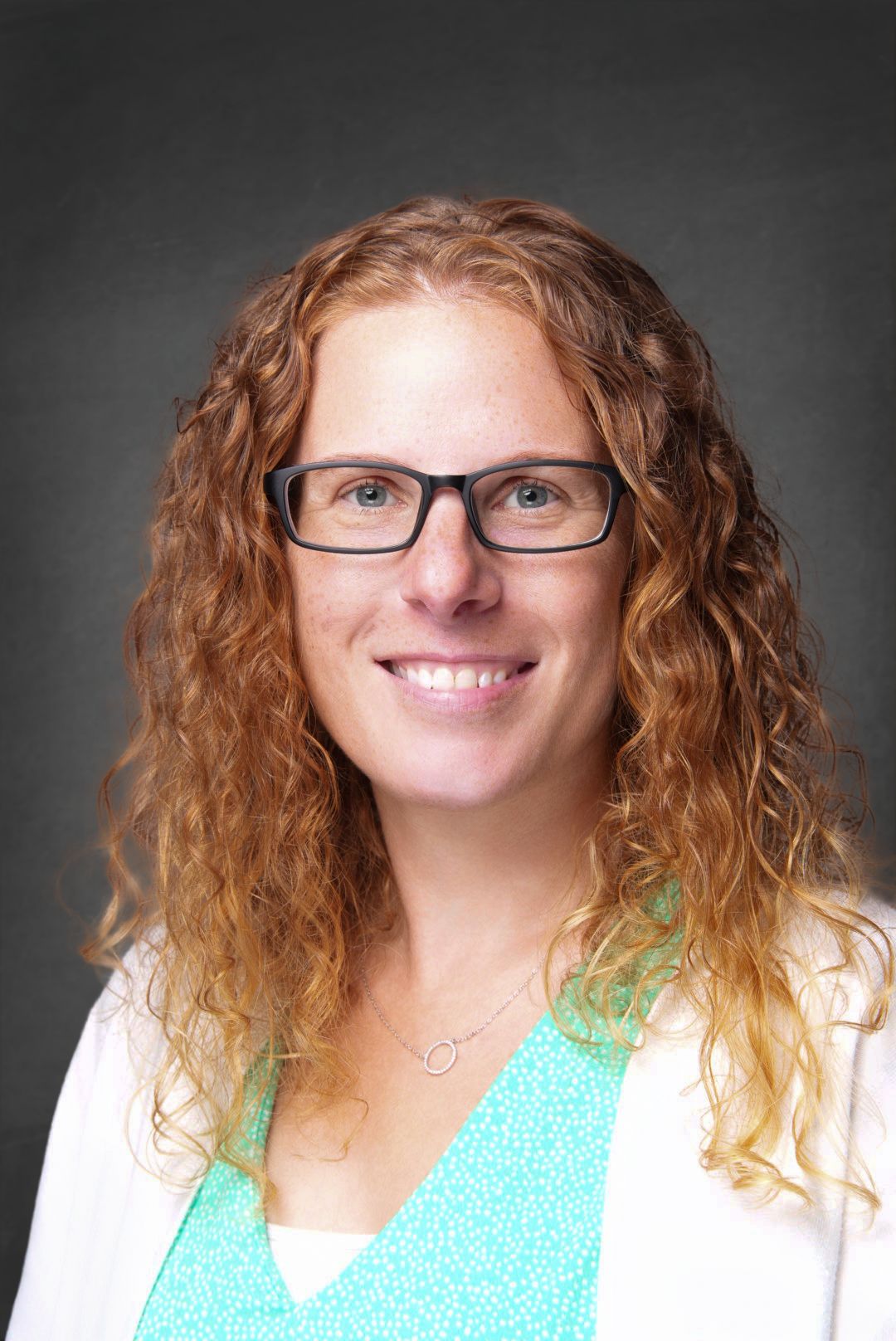 Smiling woman with long curly auburn hair and dark-framed glasses wearing a teal patterned top, white blazer and a delicate necklace.