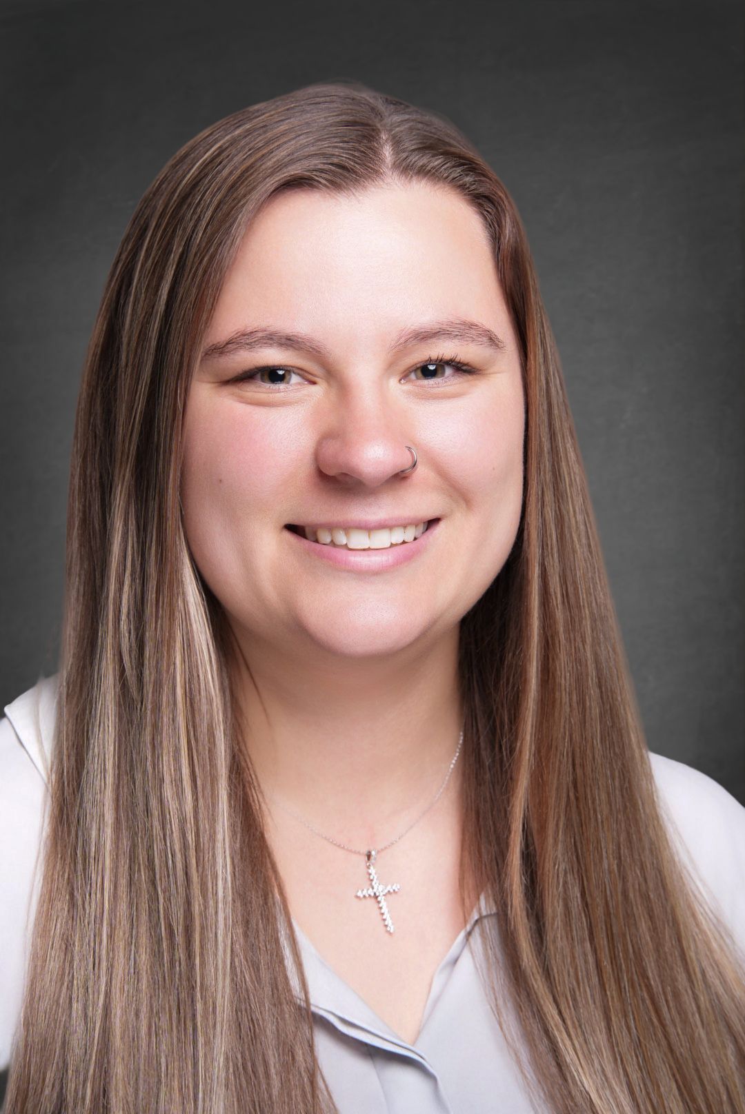 Smiling young woman with long straight brown hair with blonde highlights wearing a cross necklace and a light collared shirt in a headshot.