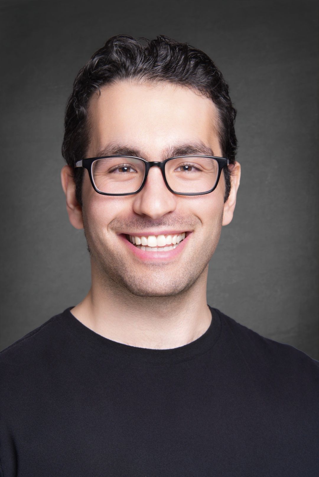 Smiling man with short dark hair and black rectangular glasses wearing a black crew neck shirt against a gray studio background.