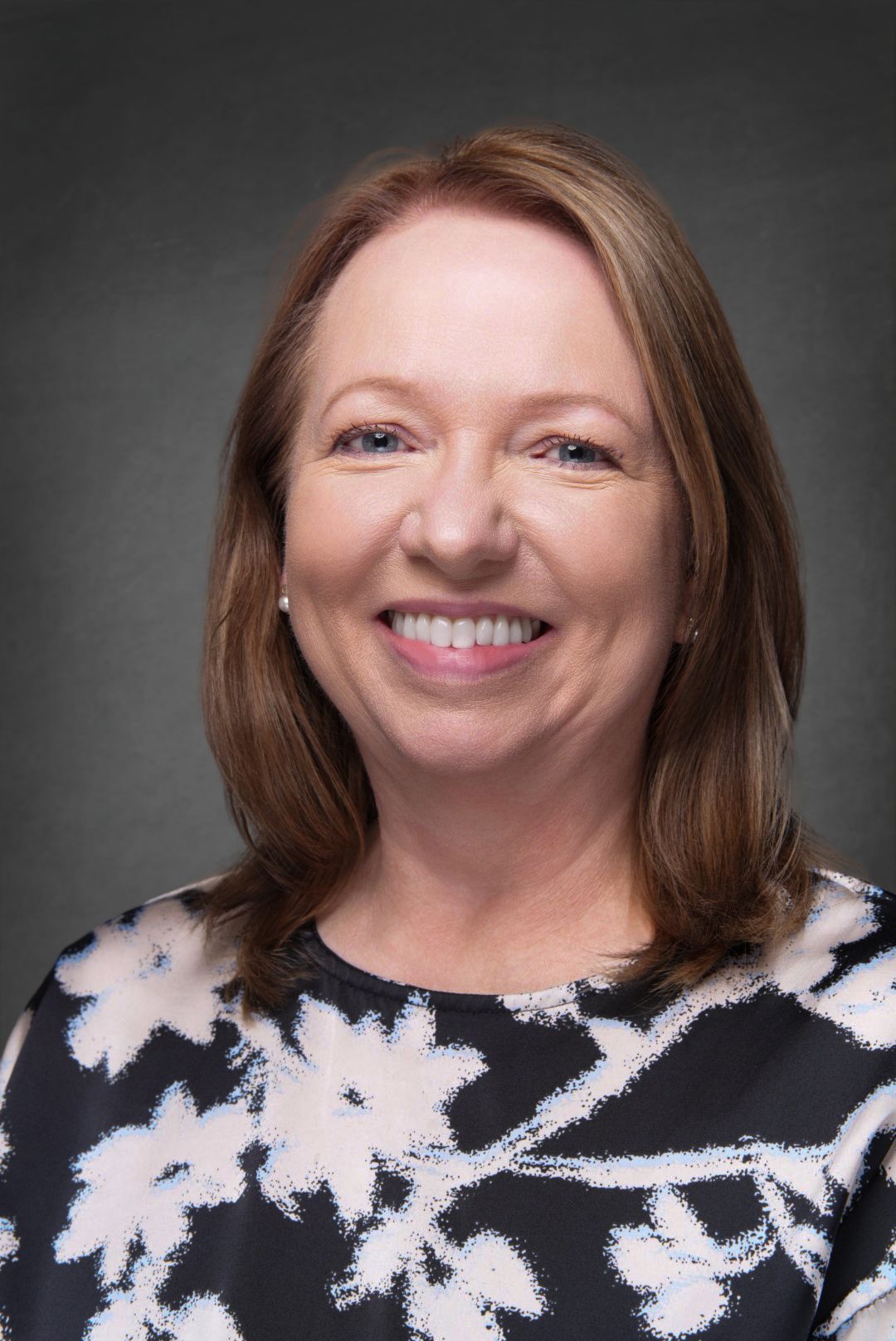 Smiling woman with shoulder-length auburn hair wearing a black blouse with white floral print, posed against a dark gray studio backdrop.