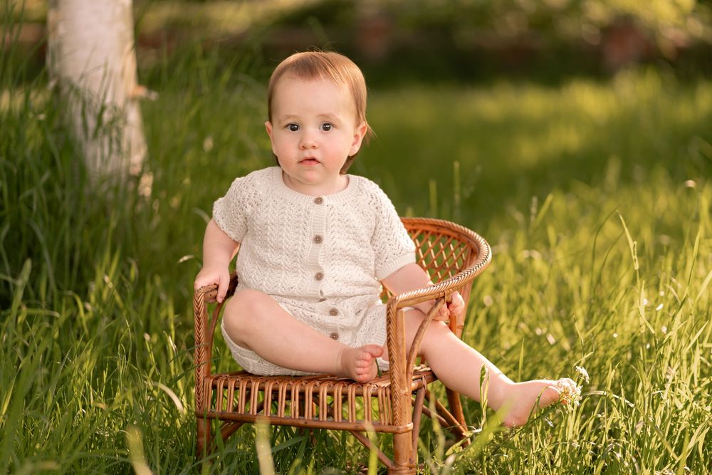 A cute baby in a white romper sits in a small wicker chair on lush green grass in warm sunlight during childhood photoshoot in Lincolnshire.