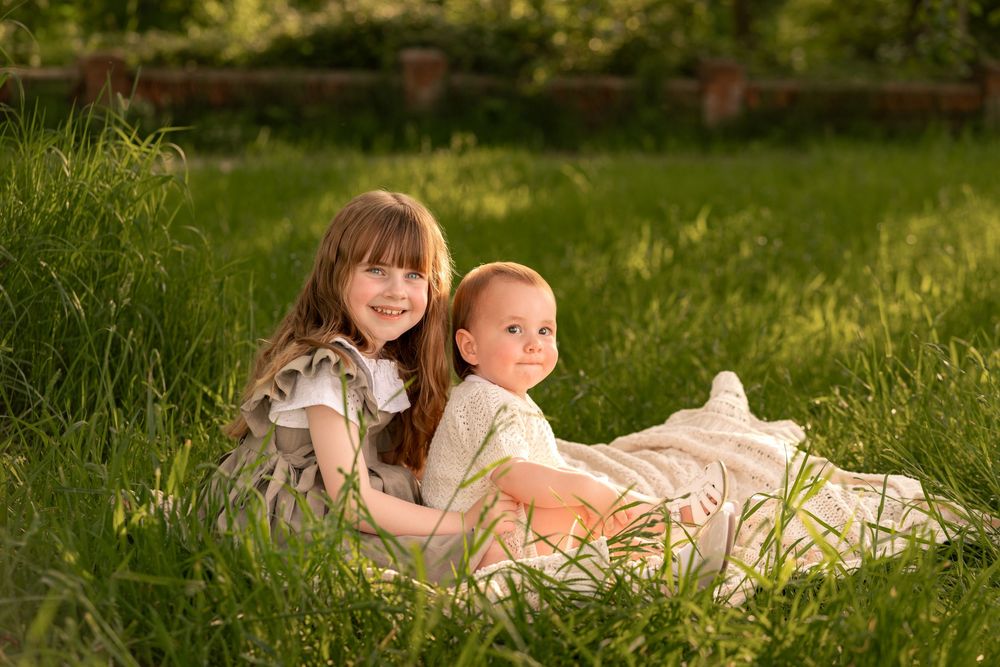 Two young siblings smile together on a blanket in lush green grass during golden hour sunlight during childhood photoshoot in Lincolnshire.