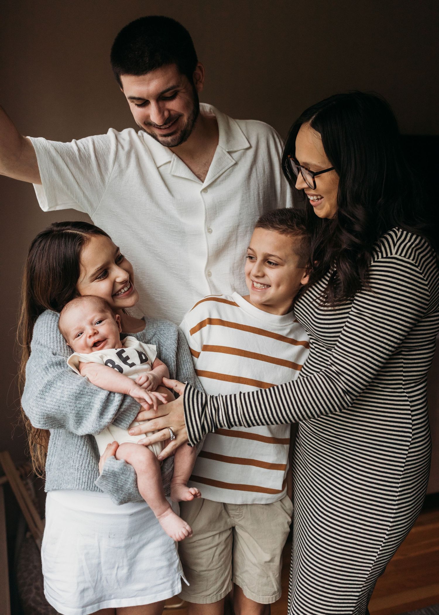 Happy family of five posing together indoors, parents smiling with three children including a newborn baby.