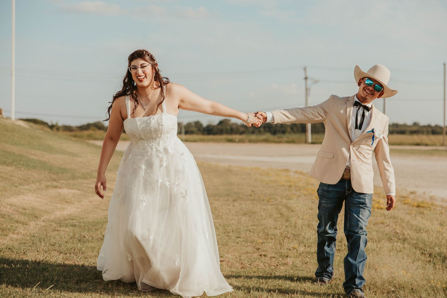 Bride in white gown holds hands with groom in cowboy hat and jeans outdoors on a sunny day.