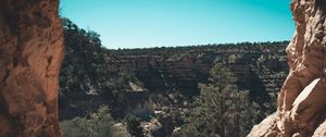 A scenic canyon landscape with red rock formations framing a view of layered cliffs and sparse vegetation under a clear blue sky.