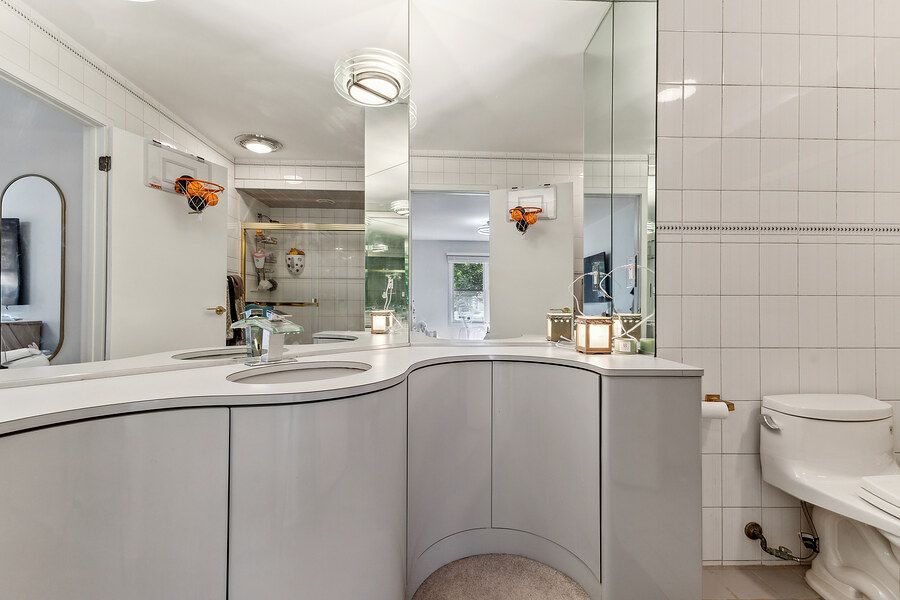 Modern bathroom with curved gray vanity, dual sinks, large mirror, and white tile walls near a bright window.