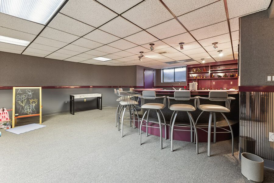 Spacious basement bar area with gray carpet, drop ceiling, bar stools, and built-in shelving perfect for entertaining.