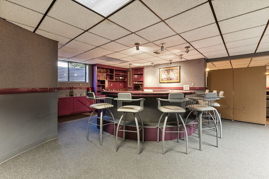 Retro basement bar area with circular counter, bar stools, drop ceiling, and built-in shelving with pink accents.