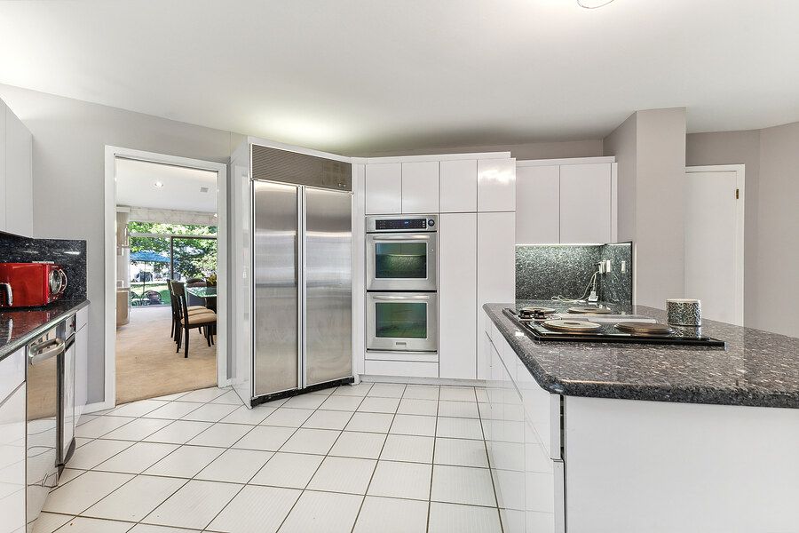 Modern white kitchen with stainless steel appliances, double wall ovens, built-in fridge, and dark granite counters.