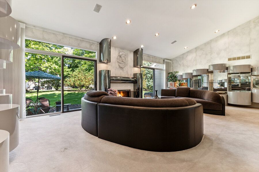 Spacious living room with curved brown sofa, high ceilings, fireplace, and floor-to-ceiling windows with garden views.