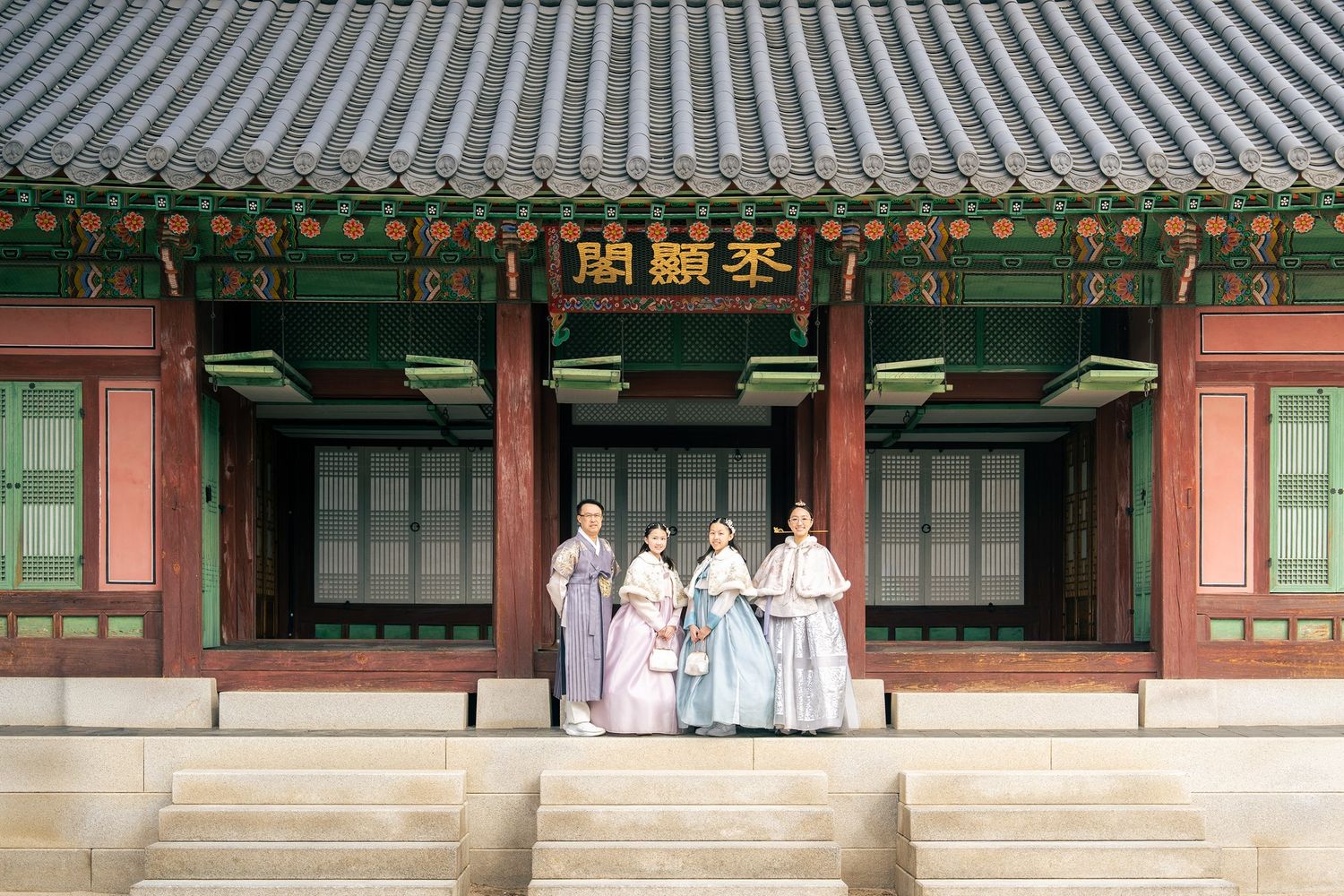 Four people in traditional Korean hanbok stand before a colorful historic palace building with ornate green and red detailing.