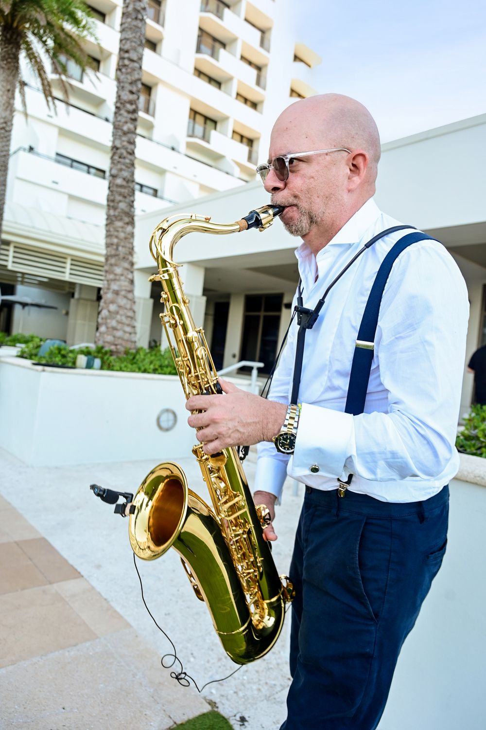 Bald man in white shirt and suspenders playing a gold saxophone outdoors near a hotel.