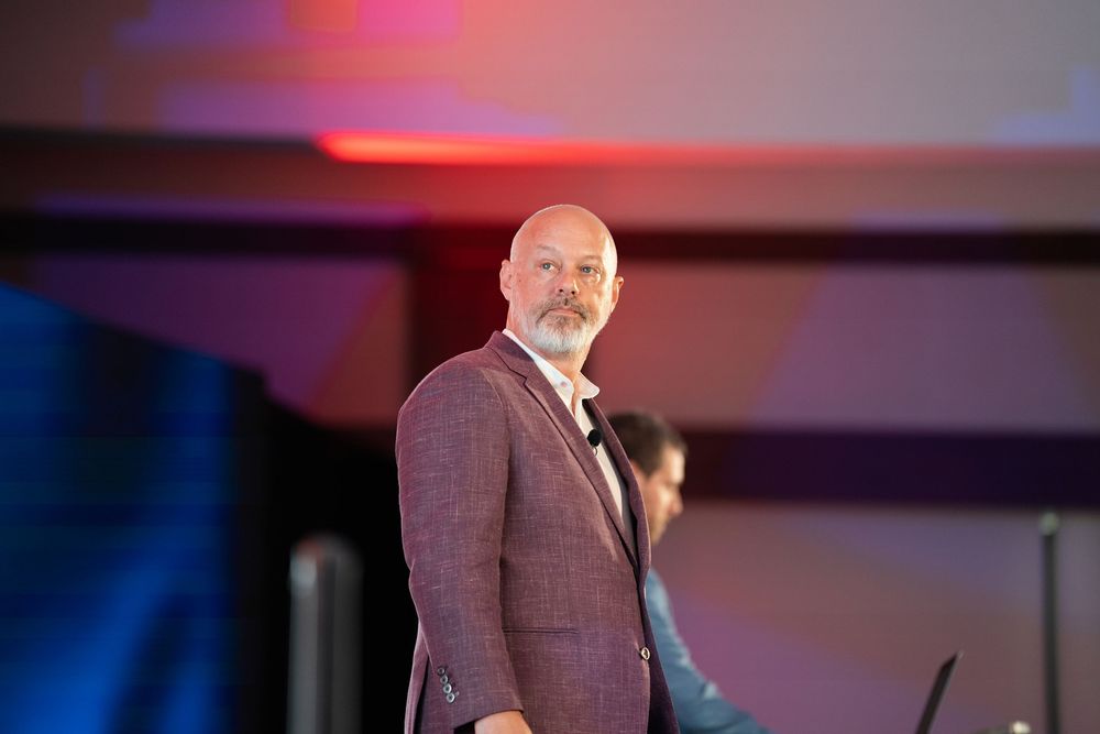 Professional speaker presents on stage at a conference wearing a brown blazer under colorful lighting.