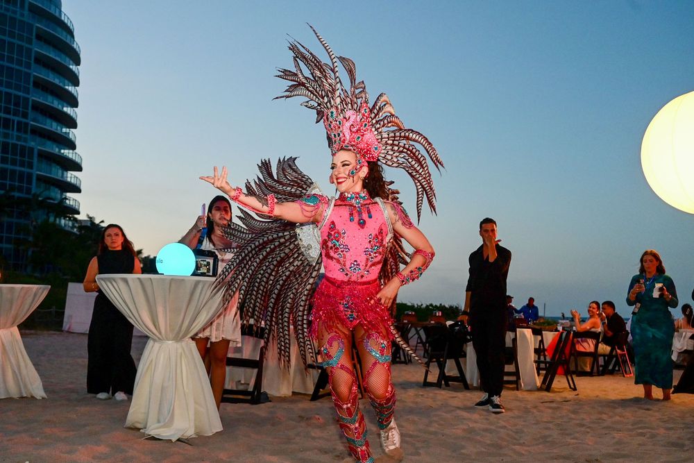 A performer in a vibrant pink costume and feathered headdress dances on a beach at dusk during an outdoor event.