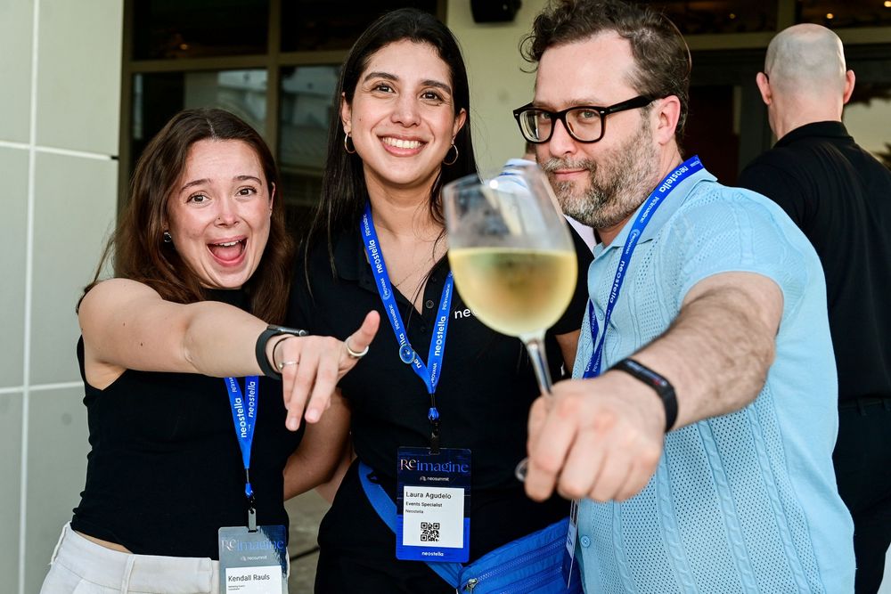 Three conference attendees smile and point at camera while holding a glass of white wine at a networking event.