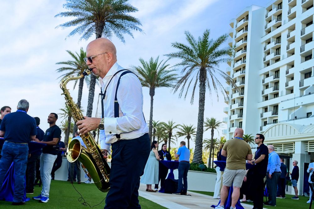Saxophonist performs outdoors at a hotel event with palm trees and guests mingling.