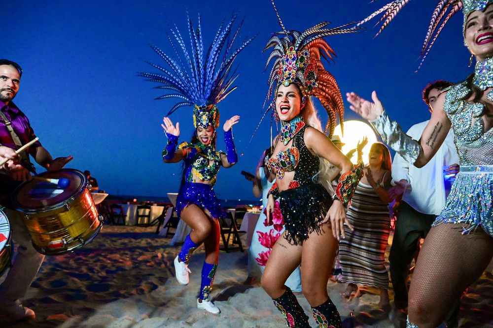 Vibrant carnival dancers in colorful feathered costumes perform on a beach at dusk with a drummer.