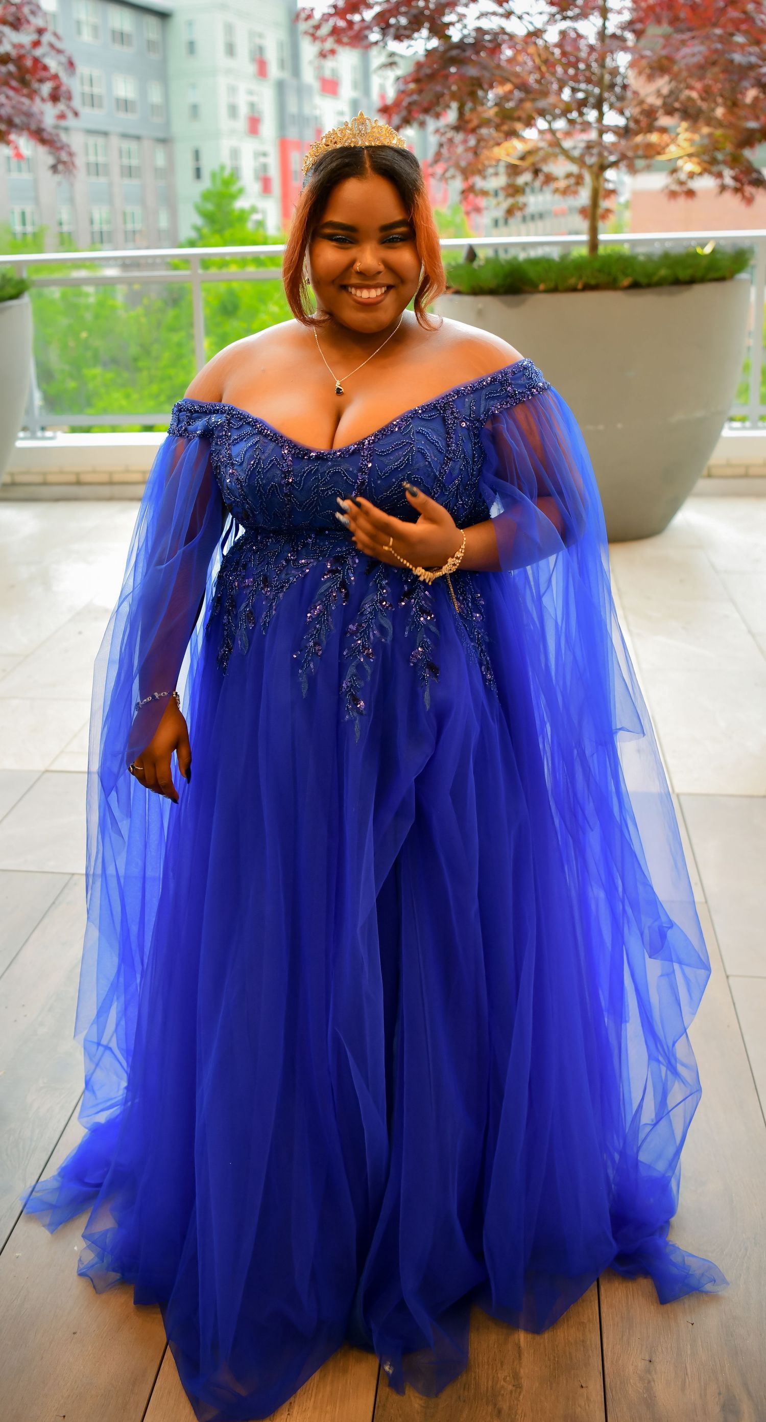 Young woman in a royal blue off-shoulder tulle ballgown with gold crown smiling at a formal event.