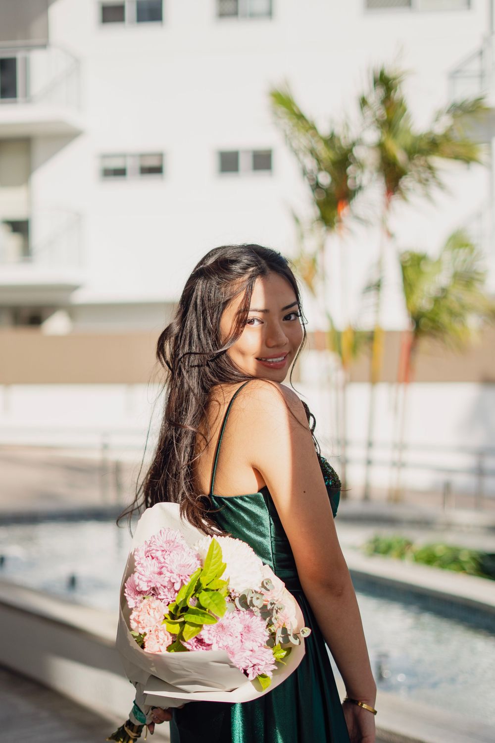Smiling young woman in elegant green dress holding a pink floral bouquet outdoors near a pool.