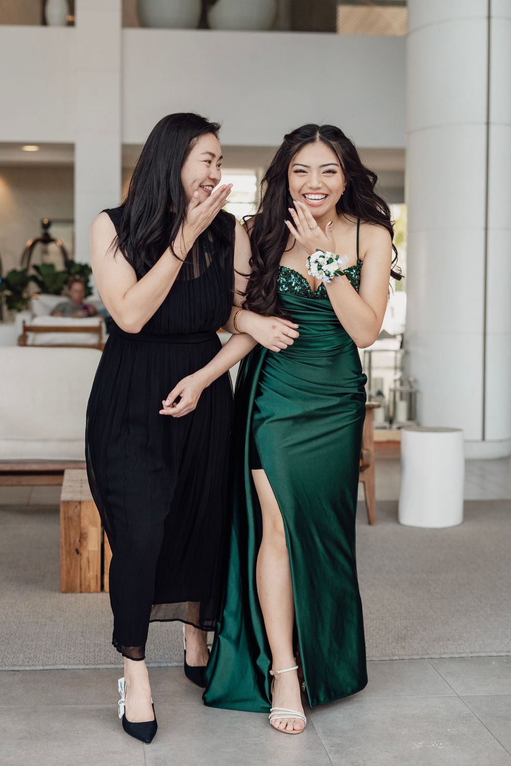 Two young women smiling together, one wearing a green satin prom gown with floral corsage.