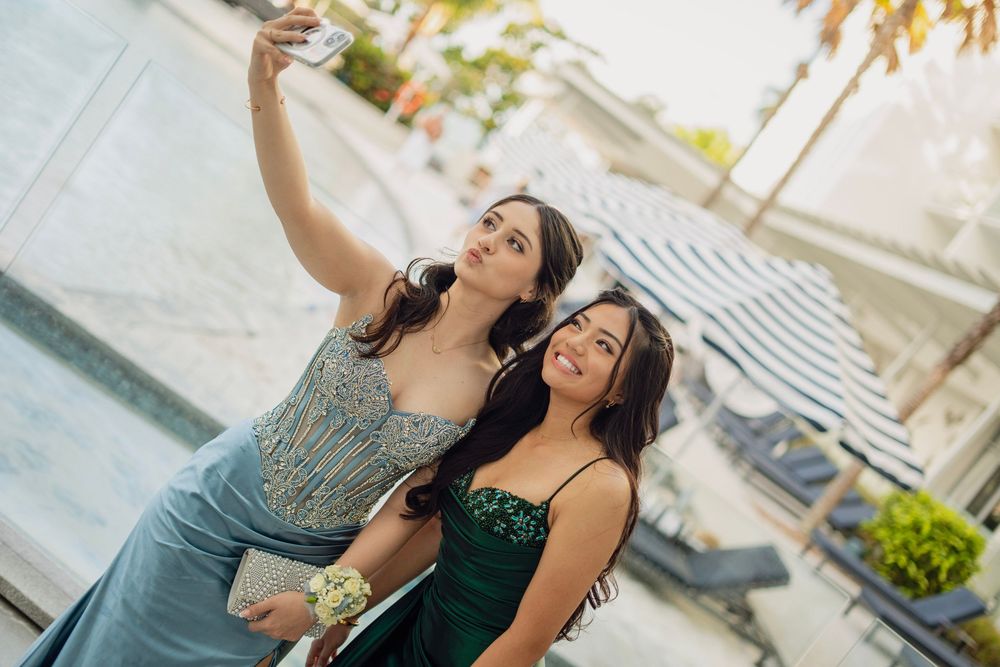 Two teenage girls in formal prom dresses take a selfie outdoors near a pool.