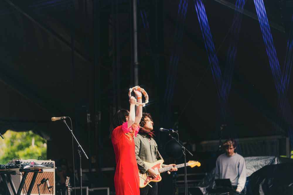 Woman in red dress raises tambourine overhead while guitarist performs on outdoor festival stage.