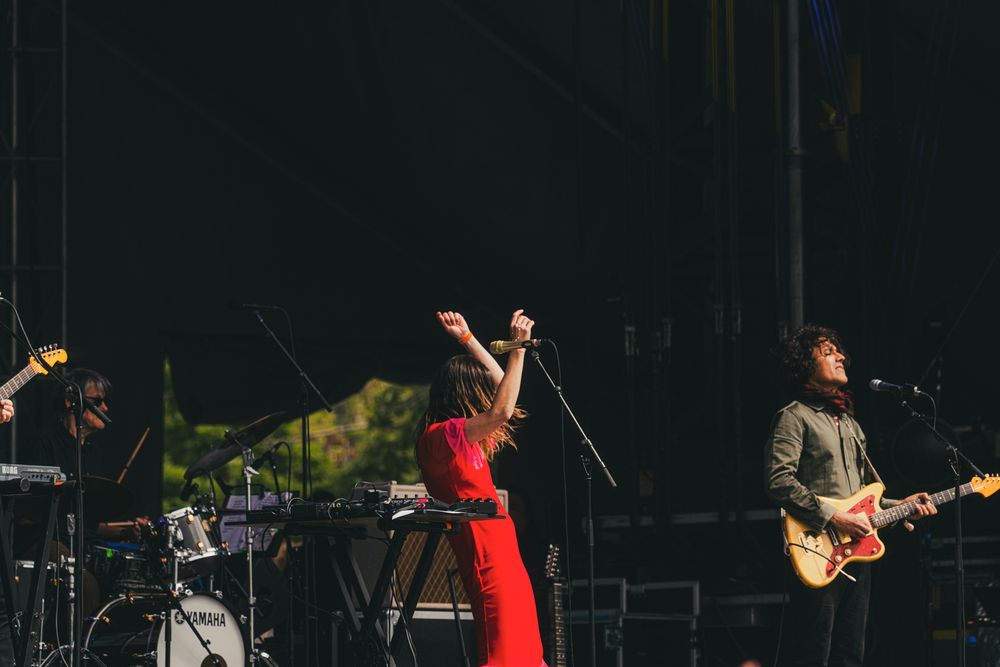 Band performing on outdoor stage, woman in red dress singing with arms raised, guitarist in suit beside her.