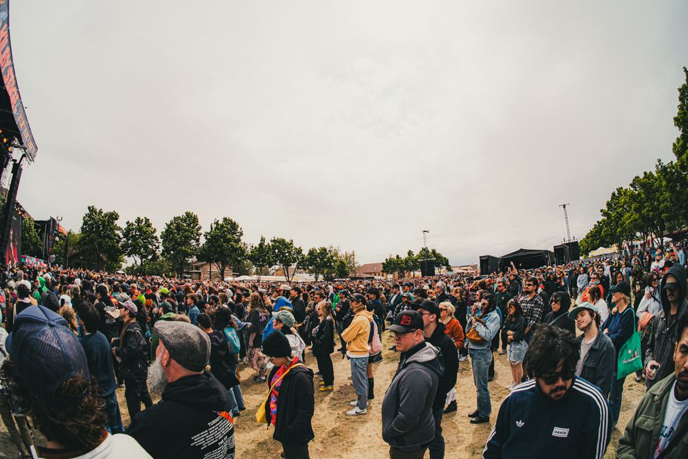 Large crowd gathered at an outdoor music festival under overcast skies, with stages and trees visible in the background.