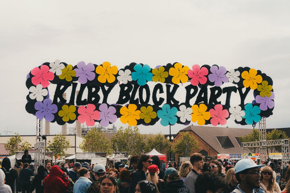 Kilby Block Party festival sign with colorful flowers on letters against a cloudy sky above a crowd.