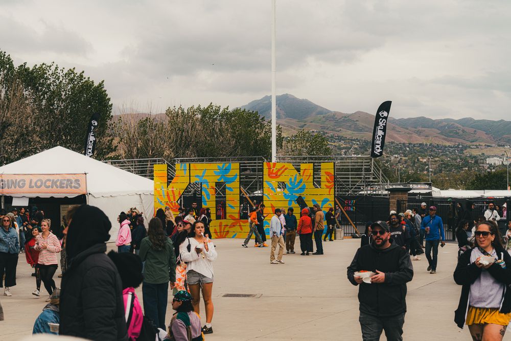 Festival crowd gathered outdoors near colorful murals with mountains in the background.