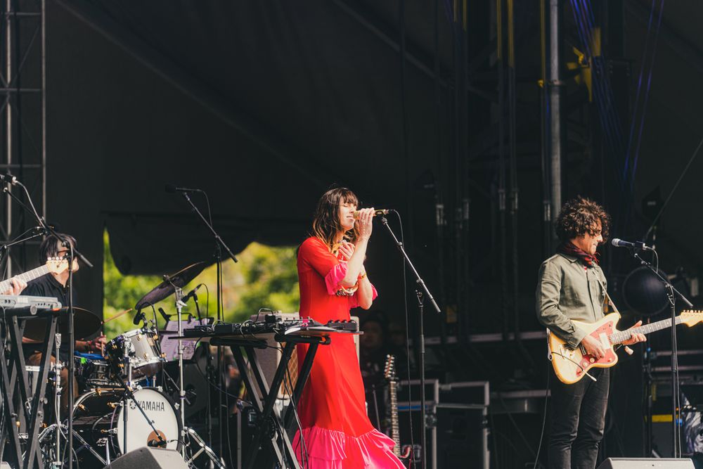 Female vocalist in red dress performs on outdoor stage with guitarist and keyboardist at live music festival.